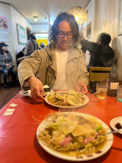 バリカタ焼きそばで有名な春来軒にて。バリカタ焼きそば2人前とおにぎり、餃子セットがテーブルに並び、髪の長い男性が嬉しそうに微笑んでいます。