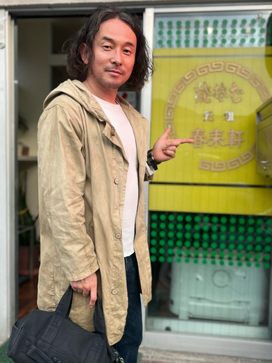 山口県のソウルフードで有名な春来軒という中華料理屋さんの前で、Cargo Shipオリジナルバッグの黒い帆布バッグCarryーOnを持つ男性がオリーブ染めしたコートを羽織りお店の前で店名を指刺しています。