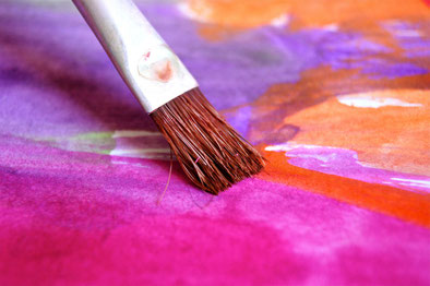 Ein schmaler, flacher Pinsel in Nahaufnahme berührt einen Aquarellblock, der mit violetten, orangefarbenen und pinken Farbtönen bemalt ist. Man sieht nur einen kleinen Ausschnitt des Aquarells.