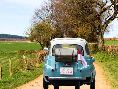 kleines blaues Auto mit weißem Dach und mit "just married" Schild und rot-weißer Flagge auf einem sandigen Feldweg von hinten aufgenommen