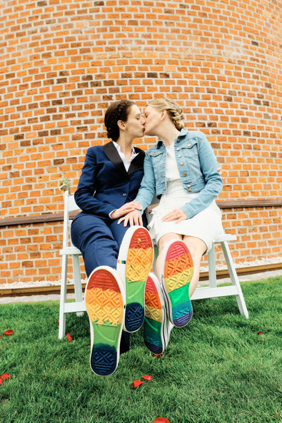 Zwei queere Bräute sitzen auf Stühlen, küssen sich und strecken ihre Schuhsohlen in die Kamera, die mit Regenbogenfarben bemalt sin.