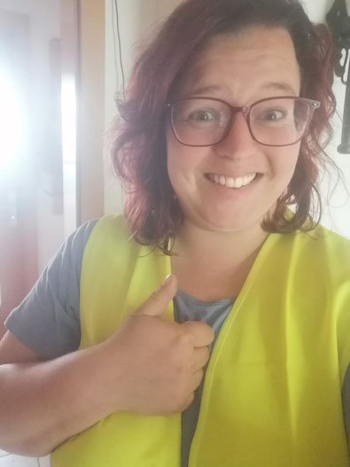 Claudia trägt ein graues T-Shirt und eine gelbe Warnweste, zeigt mit der rechten Hand einen Daumen nach oben.