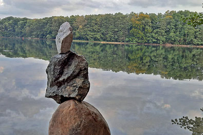 Eine Skulptur aus Steinen steht am Ufer eines großen Sees. Das gegenüberliegende Ufer ist mit Bäumen bewachsen. 