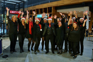 Sängerinnen und Sänger des UGC tragen Weihnachtsmützen und recken die Arme in die Luft am Flughafen BER am 23.12.2022