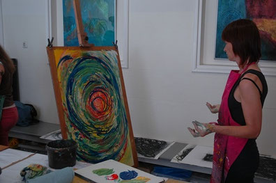 Een vrouw heeft een groot schilderij op een ezel voor haar stan. Ze heeft afstand genomen en bekijkt het beeld, met daarop een dynamische cirkel beweging van groene,blauwe en witte gebaren en met een warm oranje hart.n