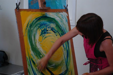 Een vrouw maakt een ruim armgebaar om een cirkel op haar schilderij te maken in groene tinten, De rest van het schilderij heeft een gele ondergrond en de rechter bovenhoek is zacht wit/blauw van tint