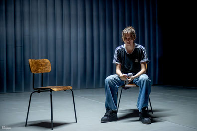 Photo de M'aime pas mort. Deux chaises se font face : une chaise vide à gauche, et Is est assise sur l'autre chaise. 