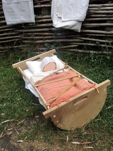 Holzwiege nach historischen Vorbild, mit schlafendem Baby und einer gestreiften Decke. Das Kind ist mit einem Ledergurt im Bettchen gesichert