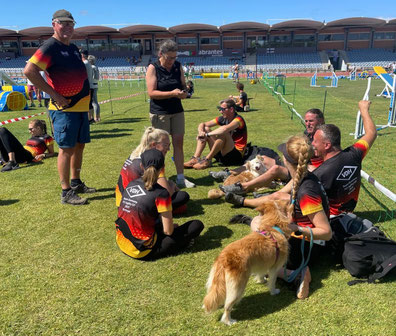 Mehrere Personen in Sportkleidung mit rot-schwarz-goldenen Trikots sitzen und stehen auf einer Sportplatzwiese, zwei Hunde sind anwesend.