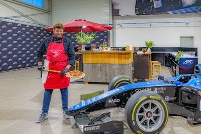 Sur cette photo on voit Walter de Mehlknepfle debout devant l'installation du Bar à Pizzas de Walter - traiteur évènementiel, tenant à la main une pelle à pizza en bois avec une pizza dessus. La scène se passe dans les ateliers de ART Grand Prix.