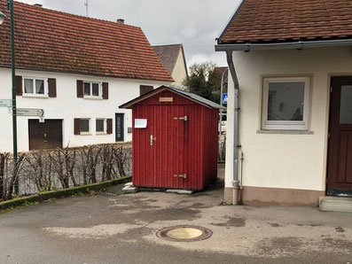 Rote kleine Holzhütte ohne Fenster, rechts daneben größeres Gebäude, links niedrige Hecke ohne Grün, davor geteerter Hof, im Hintergrund größeres Wohnhaus