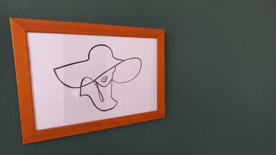Bilderrahmen aus Holz mit weißem Bild an grüner Wand. In schwarzen Linien sind die Umrisse eines Hutes und Frauengesichtes zu erkennen.