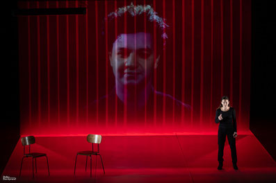 Photo du spectacle M'aime pas mort : la scène est baignée de lumière rouge, sur le rideau au lointain, on apperçoit le portrait vidéo de Sofiane. Sur l'avant de la scène, le personnage de la journaliste s'adresse au public.