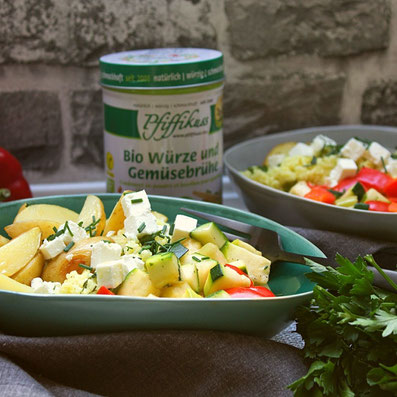 Rezept Foto Buddha Bowl mediterran, grüne Schüssel mit Kartoffel-Spalten, Zucchini und Paprika-Würfeln, Hirse und Feta, Gabel auf einer grauen Tischdecke