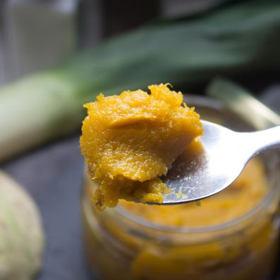 Kleiner Löffel mit orange farbener Paste, im Hintergrund ein Glas mit Paste, eine Stange Lauch und Knollensellerie