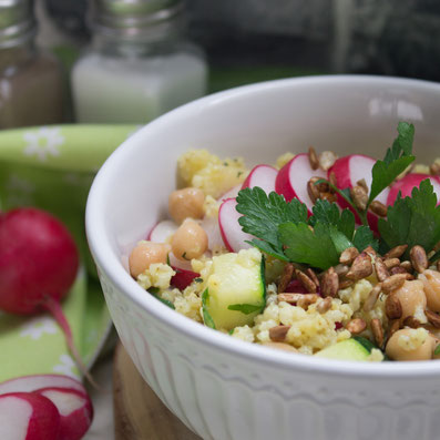 Rezeptfoto Low Carb Radieschen-Salat: Schüssel mit Hirse, Kichererbsen, Radieschen in Scheiben, Zucchini Würfel mit Petersilie. Neben der Schüssel liegt eine Zucchini und ein Radieschen mit grün.