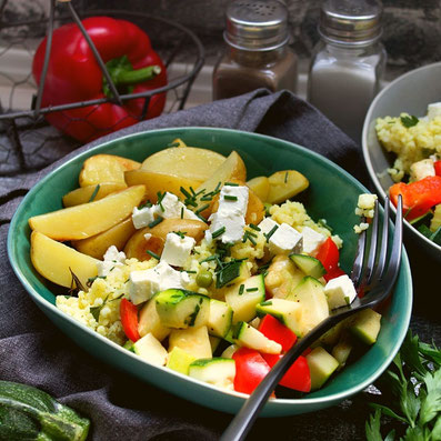 Rezept Foto Buddha Bowl mediterran, grüne Schüssel mit Kartoffel-Spalten, Zucchini und Paprika-Würfeln, Hirse und Feta, Gabel auf einer grauen Tischdecke