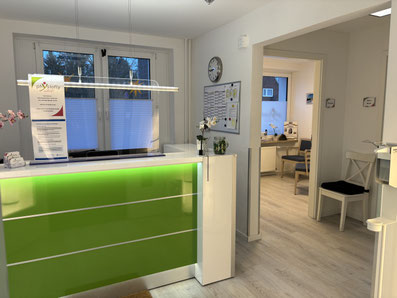 Empfangstresen mit grün beleuchtetem Frontpaneel in einer Praxis, dahinter Fenster mit halbtransparenten Jalousien, rechts Türen zu Wartezimmern mit Stühlen