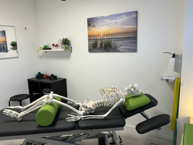 Skelettmodell liegt auf Behandlungsliege mit grüner Rolle und Kissen in einem hellen Therapieraum mit Hanteln, Spiegel und Landschaftsbild an der Wand