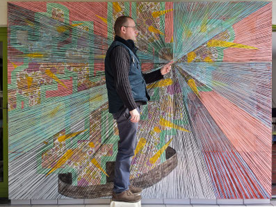 Selfie for the drawing board — artist standing in front of a large drawing board with radially stretched threads and colorful drawings, pointing toward the center.