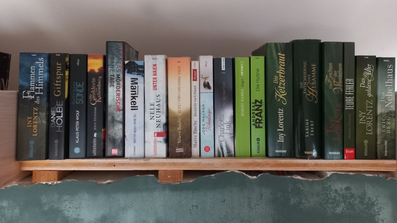 Viele Bücher stehen nebeneinander gestapelt auf einem Regal oberhalb einer grünen Wand