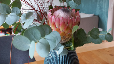 Ein Blumengesteck in einer grünen Vase steht auf einem Tisch, im Hintergrund sind ein Sofa und eine grüne Wand erkennbar.