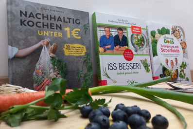 Kochbücher stehen vor Wand aufgereiht, Karotten, Blaubeeren und Frühlingszwiebel liegen auf Schneidebrett davor