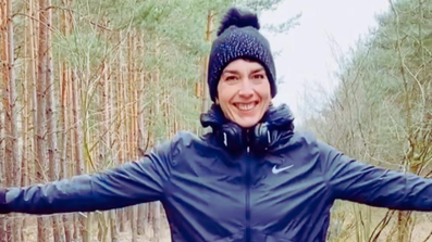 Christiane Steiger steht in blauer Sportbekleidung und Mütze auf einem Waldweg und lächelt in die Kamera, um ihrem Hals hat sie Kopfhörer.