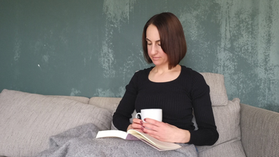 Michaela Dauscher sitzt auf einem Sofa, vor einer grünen Wand. Lächelt, ist zugedeckt und hat ein Buch auf ihrem Schoß. Sie trägt ein schwarzes Shirt, ihre Haare sind braun und schulterlang.