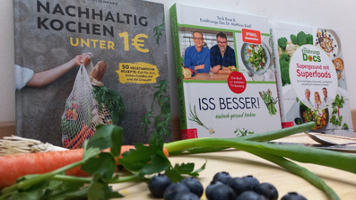 Kochbücher stehen vor Wand aufgereiht, Karotten, Blaubeeren und Frühlingszwiebel liegen auf Schneidebrett davor
