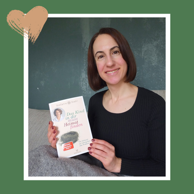 Michaela Dauscher sitzt auf einem Sofa und präsentiert das Buch das Kind in dir muss Heimat finden. Sie ist zugedeckt, lächelt, trägt ein schwarzes Oberteil, die Wand hinter ihr ist grün.
