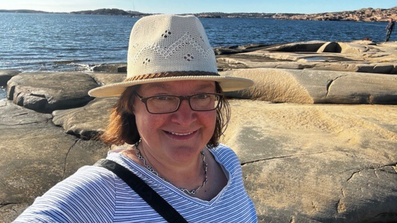 Sibille Kurdts auf felsigem Boden vor See mit Hut Brille und einem Lächeln im Gesicht