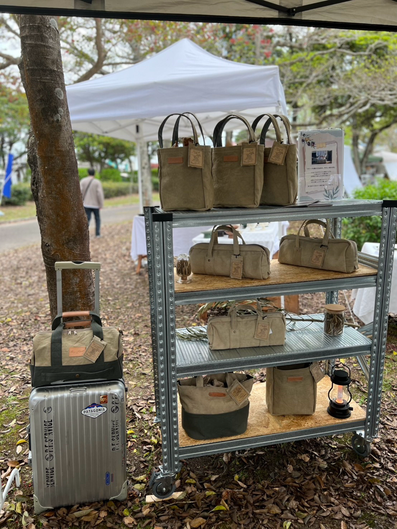 香川発、帆布バッグのお店Cargo Shipのオリジナルオリーブ染め帆布バッグ。キャリーケースの上に乗せるタイプのCarryーOnという商品とメタルシステムという自由に組み合わせできるシルバーの棚の上には、縦型トートバッグolive giraffeが３点と二段目にはオリーブ帆布バッグの横長ボストンバッグducksが２点、三段目には横長ボストンバッグolive ducksが１点と瓶に入ったオリーブの葉。そして一番下の段にはグリーンの帆布と組み合わせたミニピクニックと縦型トートバッグにバルミューダの黒いラン、