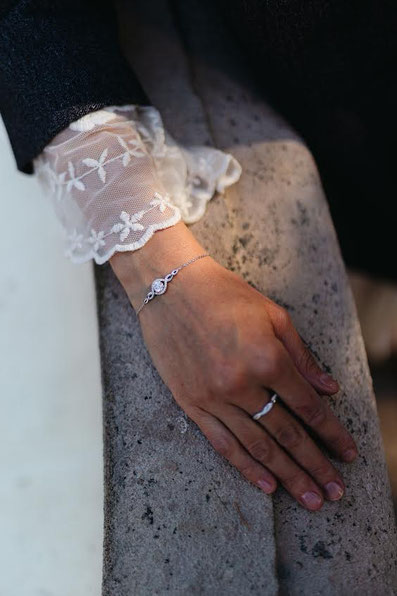 Close-up of a hand wearing a delicate silver bracelet and slim ring resting on light stone. The embroidered lace sleeve adds an elegant feminine touch.