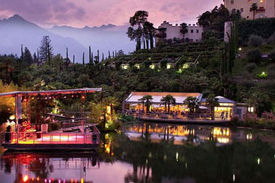 Atmosfera serale nei giardini di Trauttmansdorff con palco, lago e ristorante illuminato sullo sfondo delle montagne.