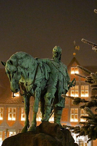 Eine Herzogsfigur aus Stein steht vor einem Perd aus Stein.  Im Hintergrund des Denkmals sieht  man die beleuchteten Fenster des Wolfenbütteler Rathauses.