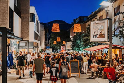 Menschen genießen einen sommerlichen Abend bei den Lana23°° Shoppingnächten in der Fußgängerzone von Lana mit Musik, Essen und Lichtern.