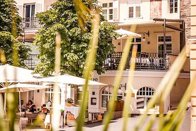 Terrazza soleggiata del ristorante Sigmund a Merano con ombrelloni bianchi, ospiti all’aperto e vista sull’elegante edificio vicino al lungofiume del Passirio