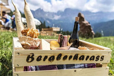 Cassa di legno con specialità altoatesine come speck, formaggio, pane, ravanelli e vino – il BerglerHarass per un picnic di gusto sullo sfondo alpino dell'Alpe di Siusi