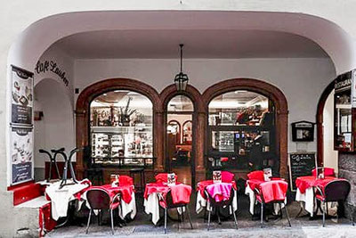 I tavolini all'aperto del caffè Lauben, situati sotto i portici di Merano, con tavoli apparecchiati di rosso, una facciata storica in legno e vista sull'accogliente sala interna del caffè.