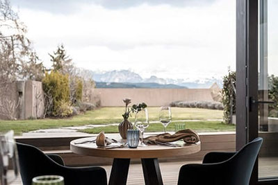 Elegant gedeckter Tisch im Restaurant Friedas im Hotel Belvedere in Jenesien mit Blick auf Garten und schneebedeckte Dolomiten – Fine Dining in exklusiver Panoramalage.