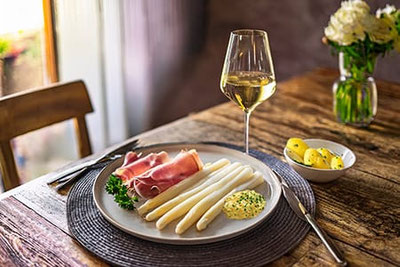Teller mit weißem Terlaner Spargel, Bozner Sauce, Südtiroler Schinken und Salzkartoffeln, serviert mit einem Glas Weißwein auf rustikalem Holztisch.