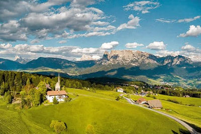 Vista panoramica dall'altopiano del Renon nei pressi di Collalbo, con l'Ansitz Kematen, i prati verdeggianti, la chiesa e le Dolomiti sullo sfondo sotto un cielo azzurro