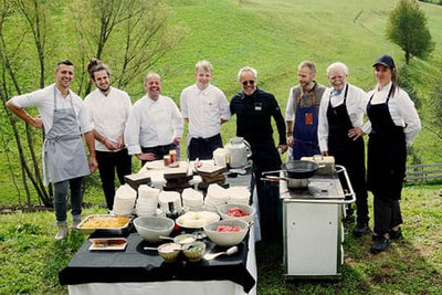I cuochi e i padroni di casa della Dispensa di Fiè sono riuniti in un prato, circondati da ingredienti freschi e postazioni di cucina, pronti per un evento all'insegna del piacere culinario.