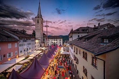 Der Kalterer Marktplatz erstrahlt bei Sonnenuntergang in festlichem Glanz. Festlich geschmückte Zelte säumen den Platz, und eine lebhafte Menschenmenge genießt die Atmosphäre.