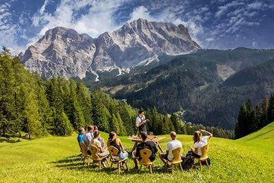 Weinverkostung im Freien in Alta Badia mit Panoramablick auf die Dolomiten, moderiert von einem Sommelier, inmitten grüner Wiesen und alpiner Bergkulisse.