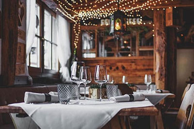 Gemütliche Stube im Restaurant Tubladel in Gröden mit rustikalen Holztischen, gedecktem Tisch, Weingläsern und stimmungsvoller Lichterkette an der Decke.