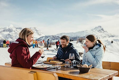 Tre persone si godono pasta e vino su un tavolo di legno in un rifugio innevato in Alta Badia durante il  skisafari, circondati da un panorama invernale soleggiato e dallo scenario delle Dolomiti.