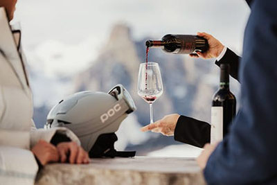 Ein Glas Rotwein wird vor winterlicher Bergkulisse eingeschenkt, ein Skihelm liegt auf dem Tisch.