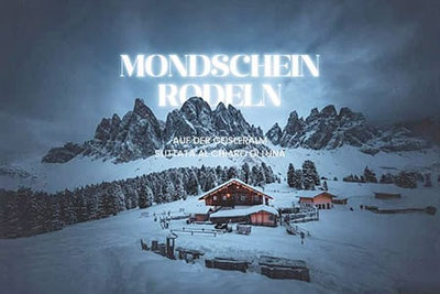 Mondscheinrodeln auf der Geisleralm in Villnöss, beleuchtete Alm vor den Geislerspitzen im Vollmond, verschneite Dolomitenlandschaft bei magischer Winterstimmung.